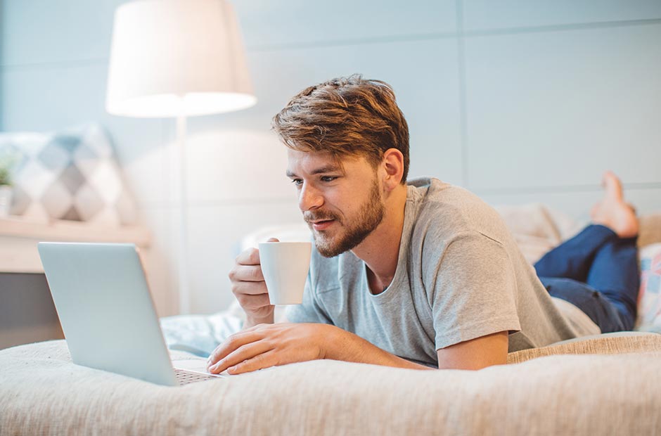 Hombre recostado en la cama, tomando una bebida caliente mientras lee sus correos electrónicos en el portátil 